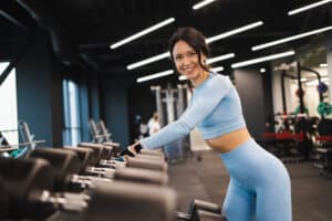 Cheerful fitness woman in blue sportswear takes dumbbells from the rack